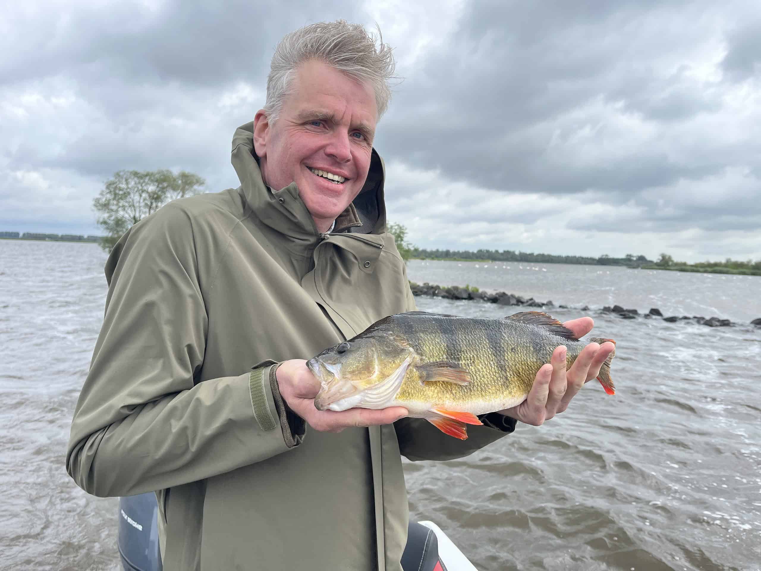 Een man in een groen jasje staat op een boot met een grote vis in zijn handen, met een bewolkte lucht en water achter hem.