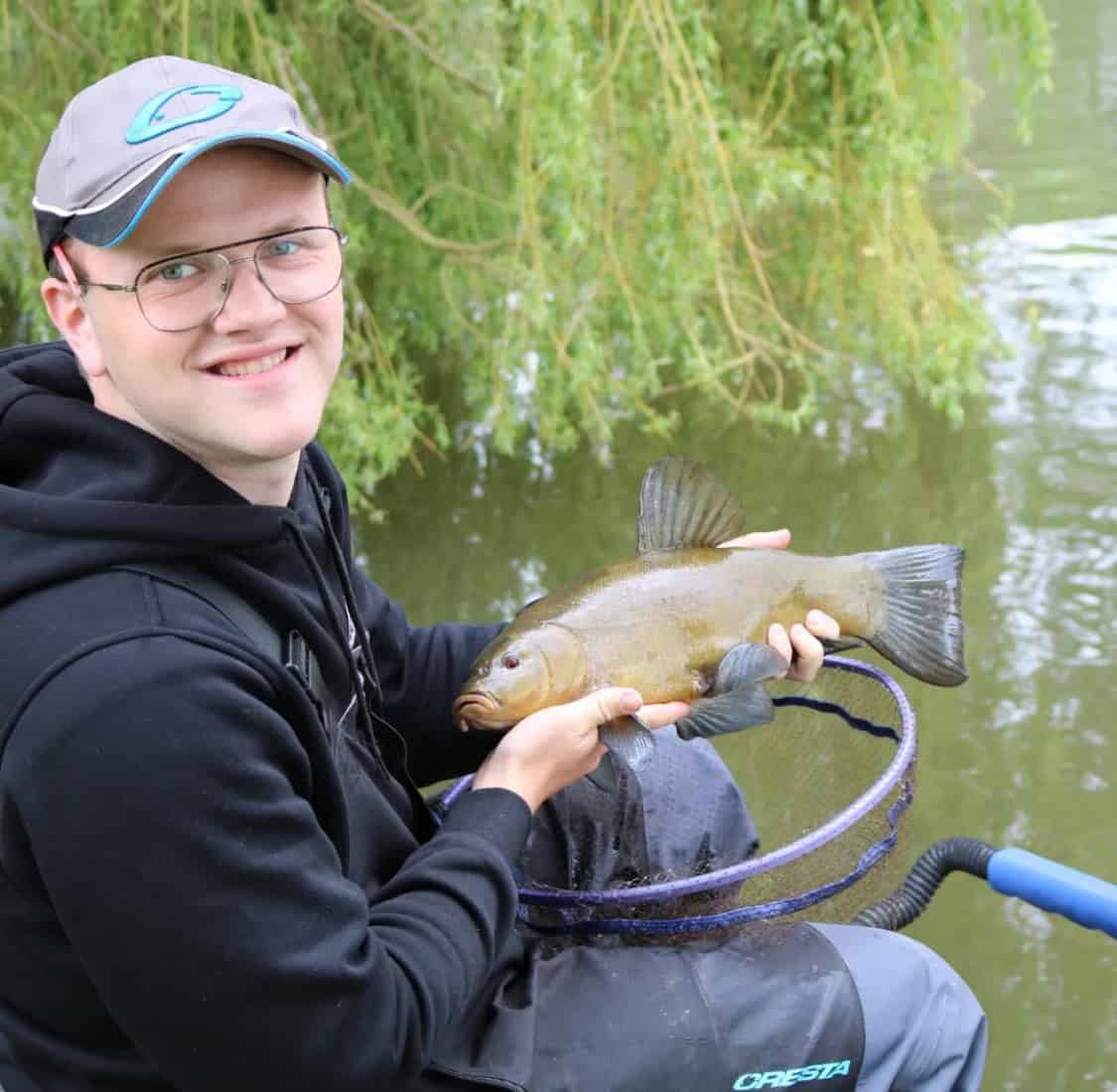 Een persoon met een bril en een pet op, zittend bij een watermassa, houdt een gevangen vis boven een net. Vlakbij staat een busje geparkeerd tegen de achtergrond van weelderig groen gebladerte.