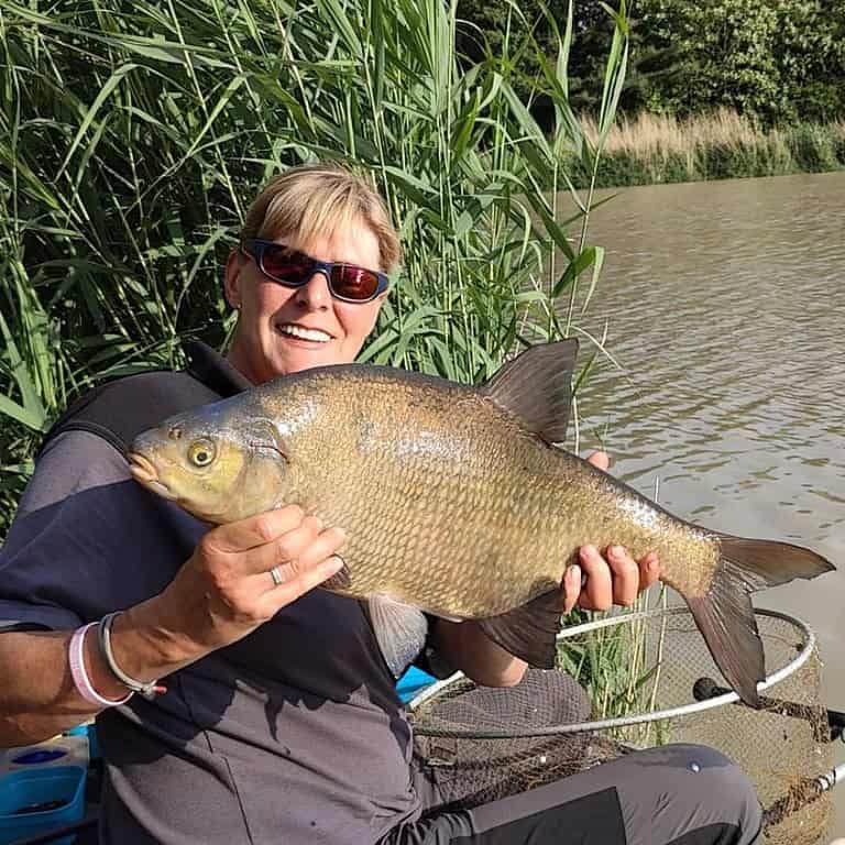 Persoon met een zonnebril en een donker shirt die een grote vis vasthoudt voor hoog riet bij een watermassa, onderdeel van onze kleine editie Vangsten van de week.