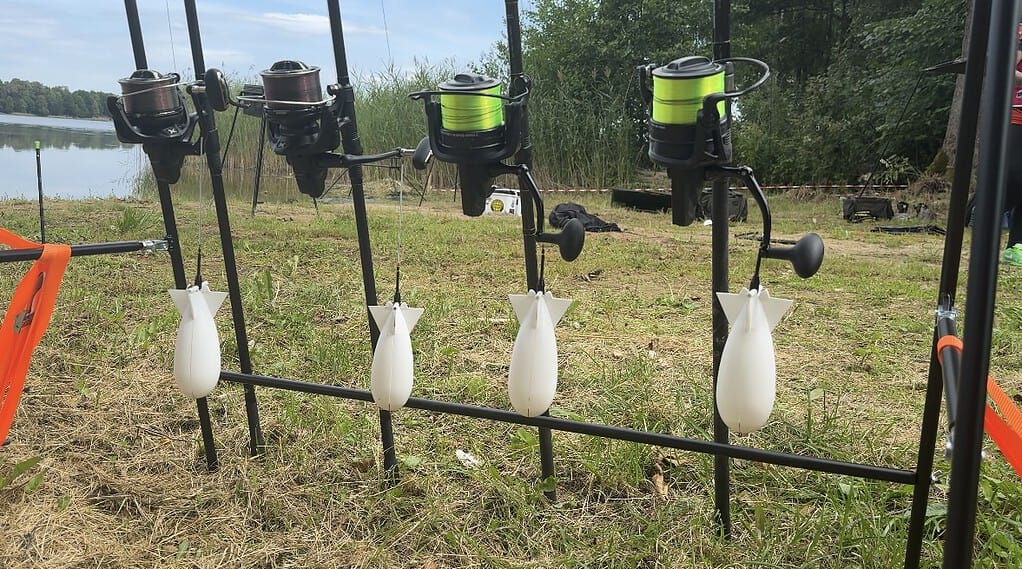 Bij een meer staan vier hengels met groene en bruine molens en vier witte raketvormige aashouders opgesteld, die naadloos opgaan in het natuurlijke landschap van gras en bomen, een belichaming van automatische concepten.