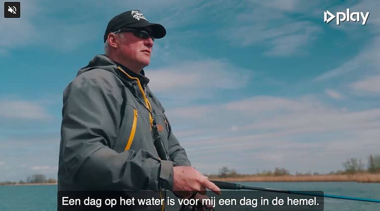 Een persoon met een pet en een reflecterende zonnebril is aan het vissen op een watermassa, met een blauwe lucht en enkele wolken op de achtergrond. Onderaan zijn Nederlandse ondertitels zichtbaar.