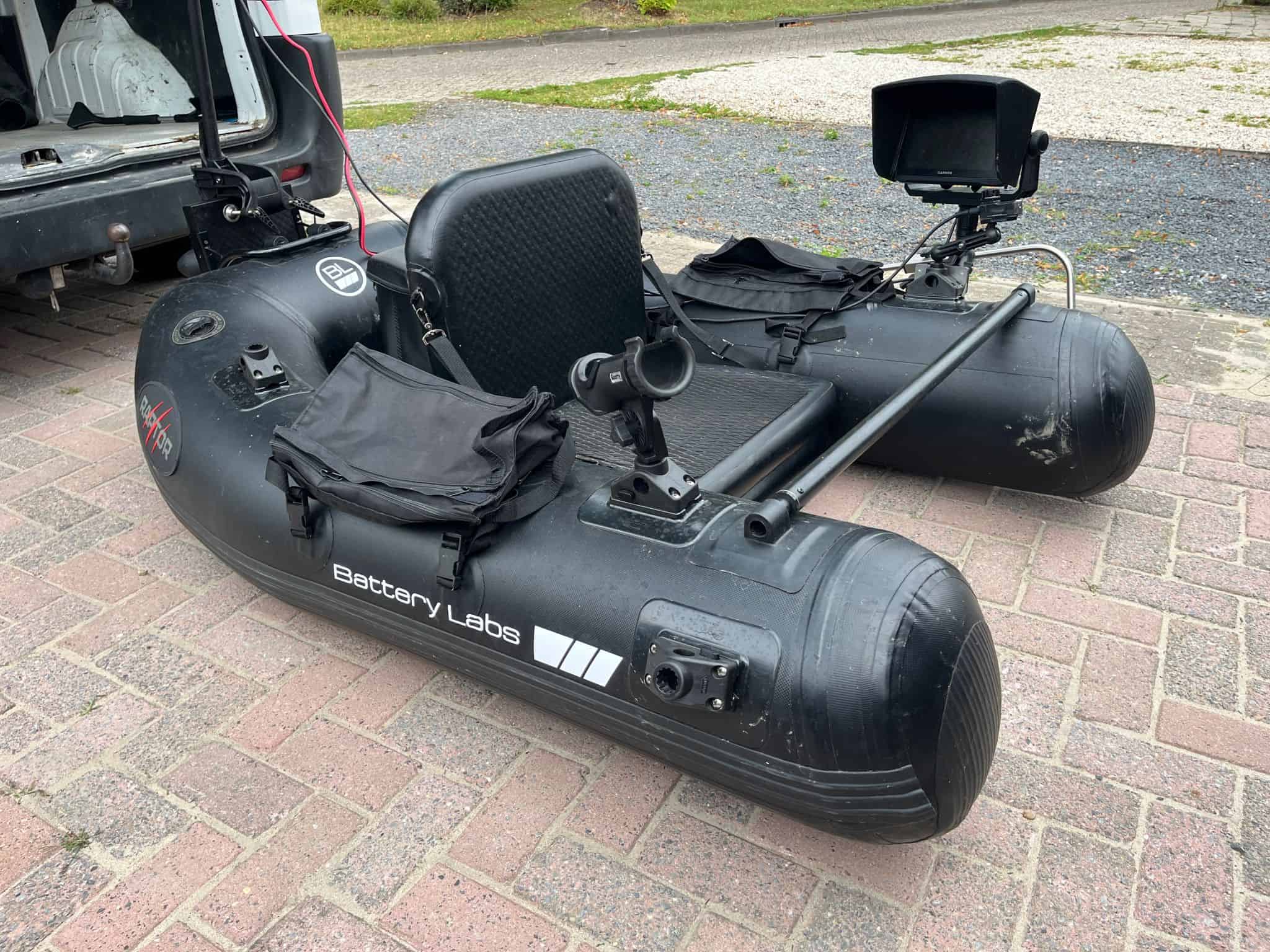 Een opblaasbare zwarte boot met het label "Battery Labs" met gemonteerde apparatuur en een open achtercompartiment, perfect voor de eerste inspanningen van een bellyboatvisser. De boot staat op een met stenen geplaveide ondergrond naast een voertuig.