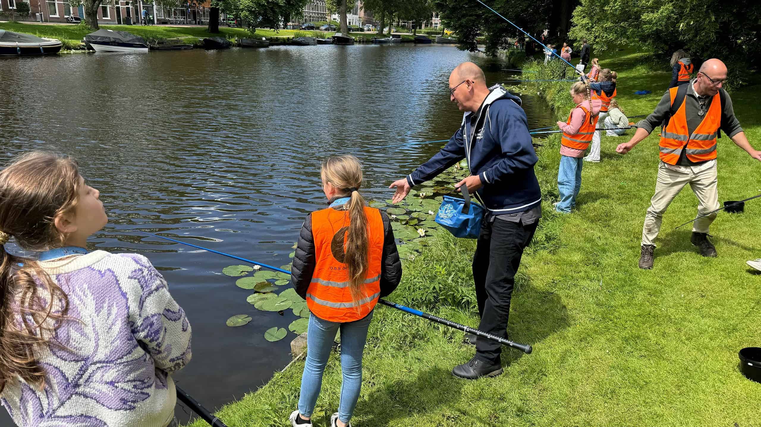 Een groep mensen, bestaande uit kinderen en volwassenen, draagt oranje hesjes en is aan het vissen aan de oever van een kalme rivier op een grasveld in een parkachtige omgeving. Ze vangen de essentie van conceptvorming te midden van de rust van de natuur.