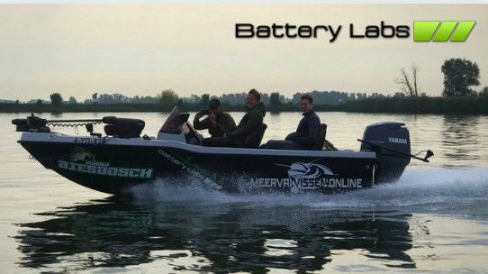 Drie personen zitten in een zwarte motorboot met "Battery Labs" en andere logo's op een kalm waterlichaam, één persoon bestuurt en twee zitten erachter. De achtergrond toont een boomgrens en een bewolkte lucht. Dit Nederlandse topmerk vaartuig is uitgerust met geavanceerde stroomvoorziening voor ononderbroken prestaties.