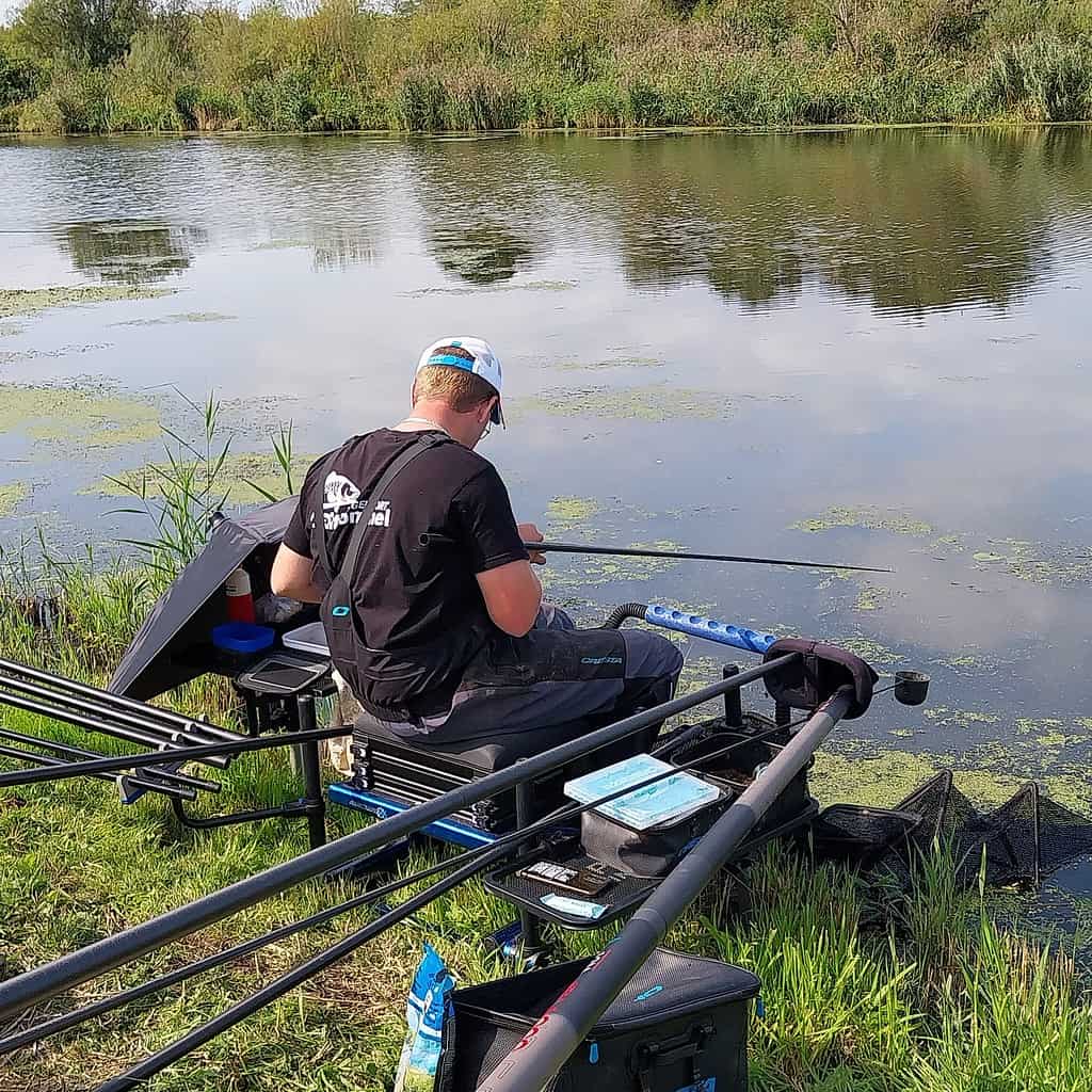 Een persoon met een zwart shirt en pet zit bij een kalm meer, visgerei ordenend, met verschillende hengels en een tacklebox in de buurt. Het meer is gedeeltelijk bedekt met algen.