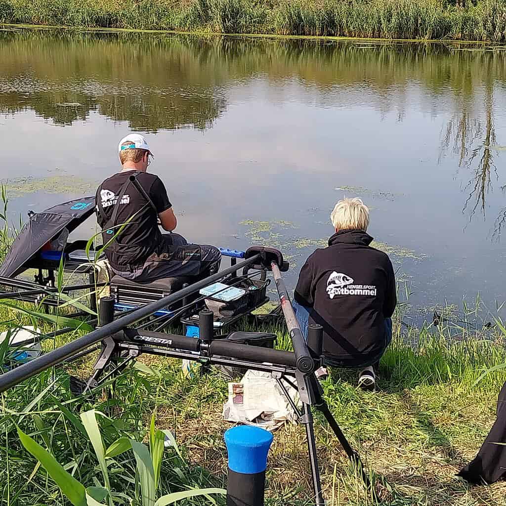 Twee mensen in bijpassende zwarte shirts en hoeden, zittend bij een meer met visgerei. Het water is kalm en er is groen rondom de scène.