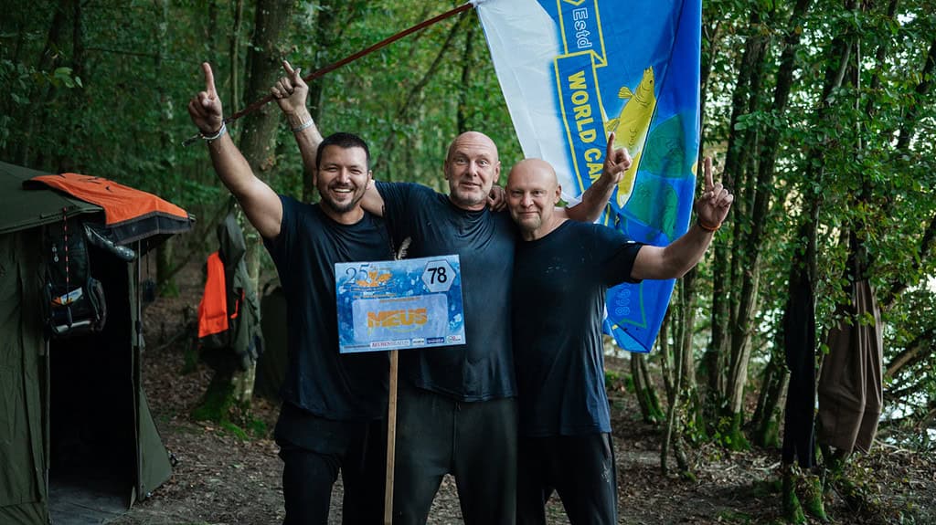 Drie mannen vieren feest in een bosgebied, met een bord waarop "78" staat en een vlag waarop "World Carp Classic 2024" staat. Ze lachen, met opgeheven armen in de overwinning bij Lac de Madine. Tenten en uitrusting zijn zichtbaar op de achtergrond.