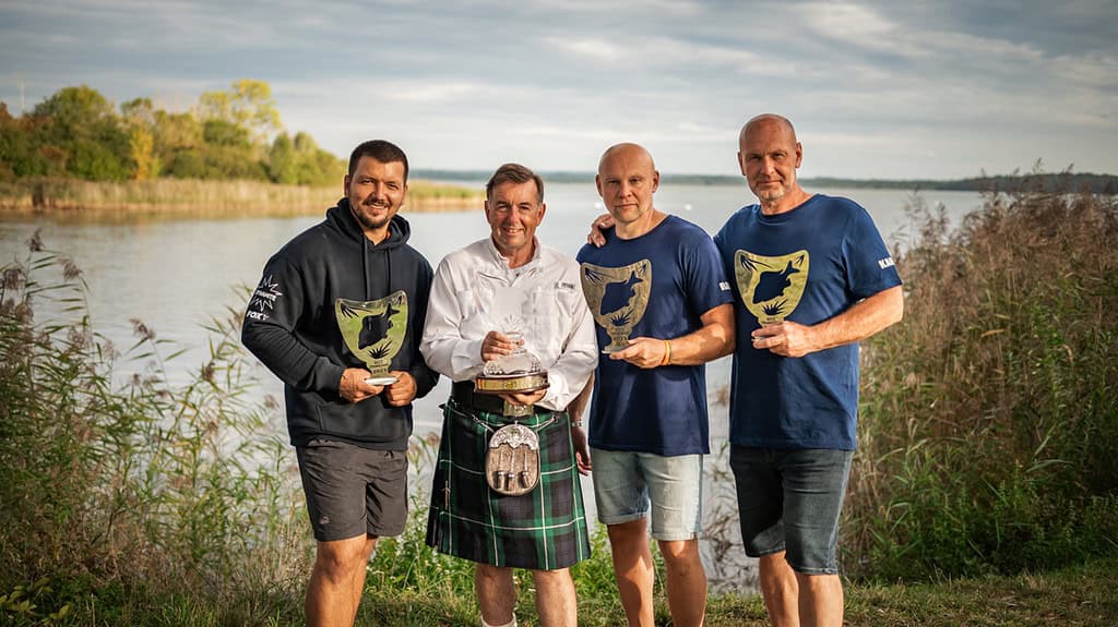 Vier mannen staan buiten bij een meer, waarschijnlijk Lac de Madine in Frankrijk. Drie dragen bijpassende blauwe shirts met een logo, terwijl één gekleed is als chef-kok. Ze tonen trots verschillende onderscheidingen. Bomen en riet vormen de serene achtergrond, die het decor vormt voor wat onderdeel zou kunnen zijn van de World Carp Classic-festiviteiten.