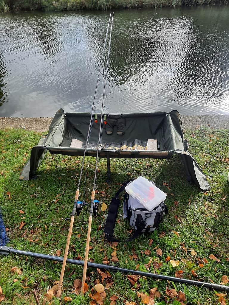 Twee hengels rusten op een standaard aan een rivier, met een tacklebox en een tas op het gras ernaast. Krijn van Urk legt dit geslaagde tafereel perfect vast in zijn nieuwste doodaassessie.