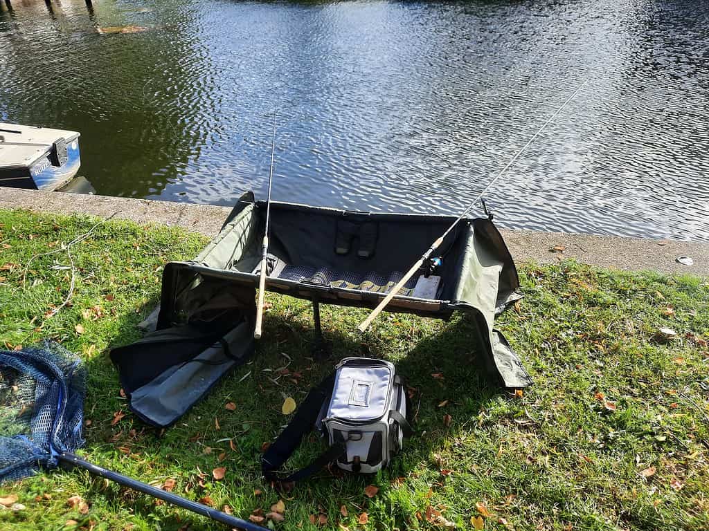 Een visuitrusting langs de rivier met twee hengels op een standaard, een open vistas op het gras, een schepnet en een groene stoel vormen het decor voor wat een succesvolle doodaassessie belooft te worden.