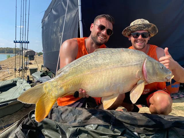 Twee mannen van het Nationaal Karperteam houden buiten een grote vis vast terwijl ze lachend naar de camera kijken. Visgerei en een watermassa zijn op de achtergrond zichtbaar, perfect vastgelegd door Media Manager Cindy Horstman.