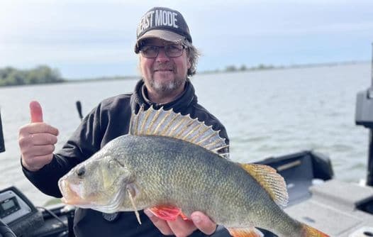 Een persoon met een "FAST MODE"-hoed houdt een grote vis vast met één hand en steekt met de andere hand zijn duim omhoog terwijl hij op een boot in het water staat en viert de succesvolle vangsten die hij dit najaar is gaan zien.