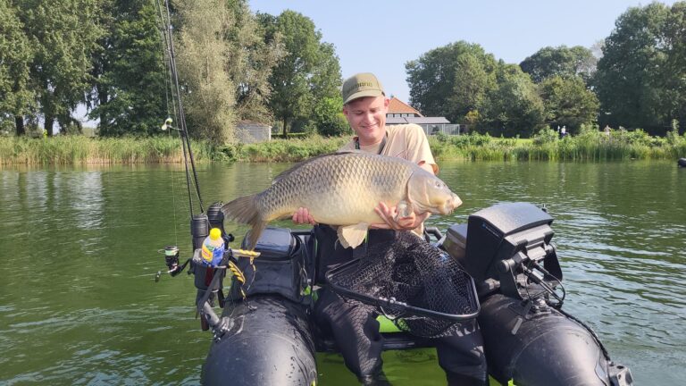 Een persoon in een kleine opblaasbare bellyboat, met een grote vis in zijn armen, bevindt zich op een kalm meer met bomen en huizen op de achtergrond. Hij doet waarschijnlijk mee aan een sportvisserijwedstrijd.