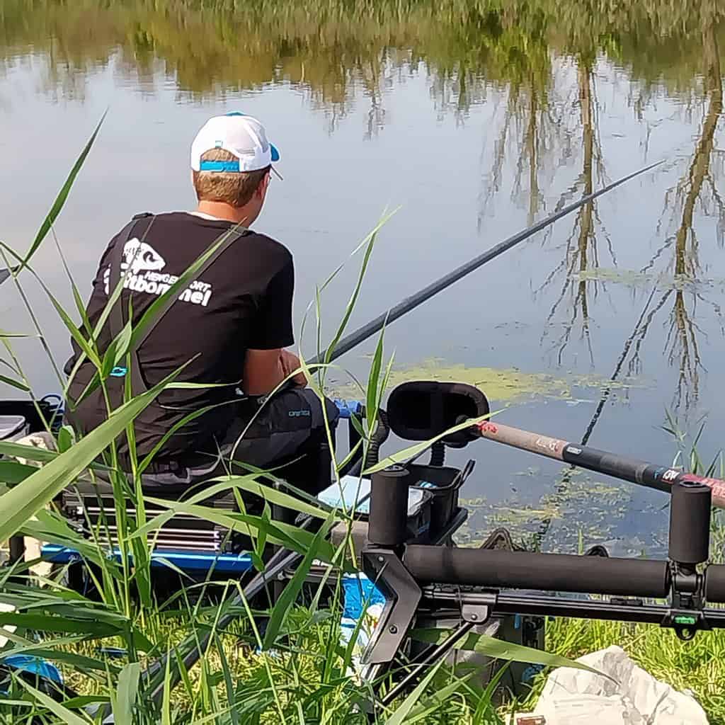 Een persoon met een blauw-witte pet en een zwart shirt zit op een krukje in het hoge gras te vissen in een vijver, met zijn gezicht van de camera af.