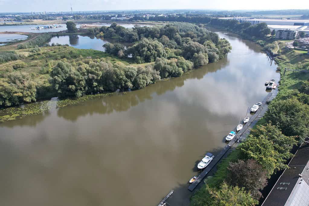 Luchtfoto van een rivier met bomen en kleine boten, mogelijk aan het vissen. In de buurt is een woonwijk en uitgestrekte begroeiing onder een gedeeltelijk bewolkte lucht, wat doet denken aan scènes die Paul Breems heeft vastgelegd.