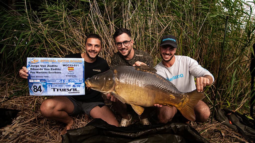 Drie mannen knielen in het hoge gras bij Lac de Madine, met trots een grote vis en een bord met details over hun indrukwekkende vangst voor de De World Carp Classic van 2024. Ze lachen recht in de camera.