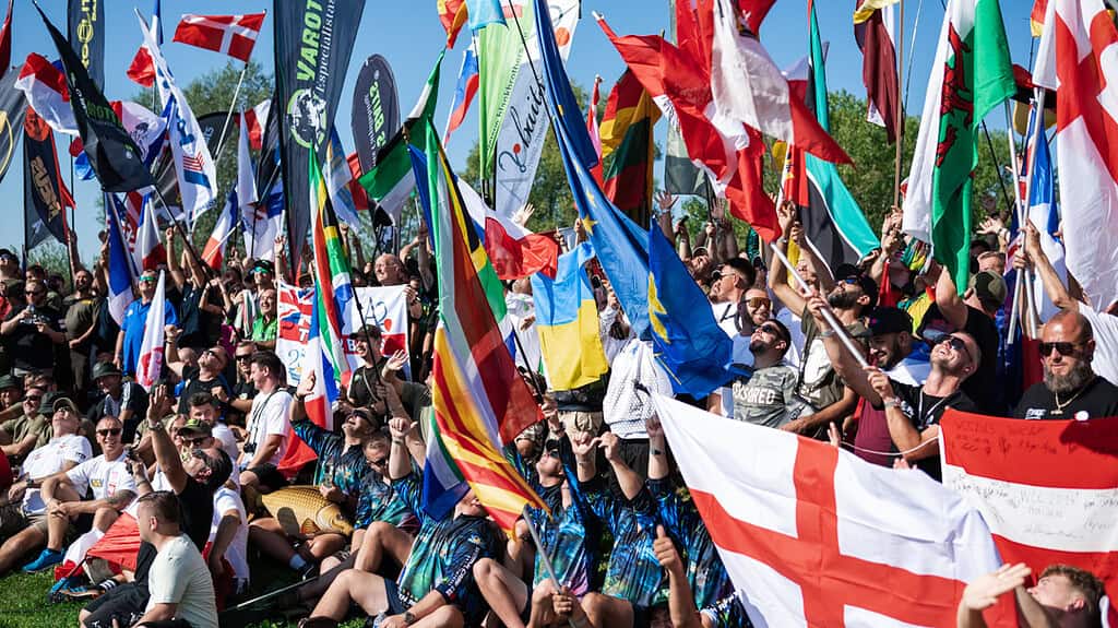 Een grote menigte mensen, sommigen met vlaggen van verschillende landen, verzamelde zich op een zonnige dag buiten bij de De World Carp Classic 2024 in Lac de Madine.