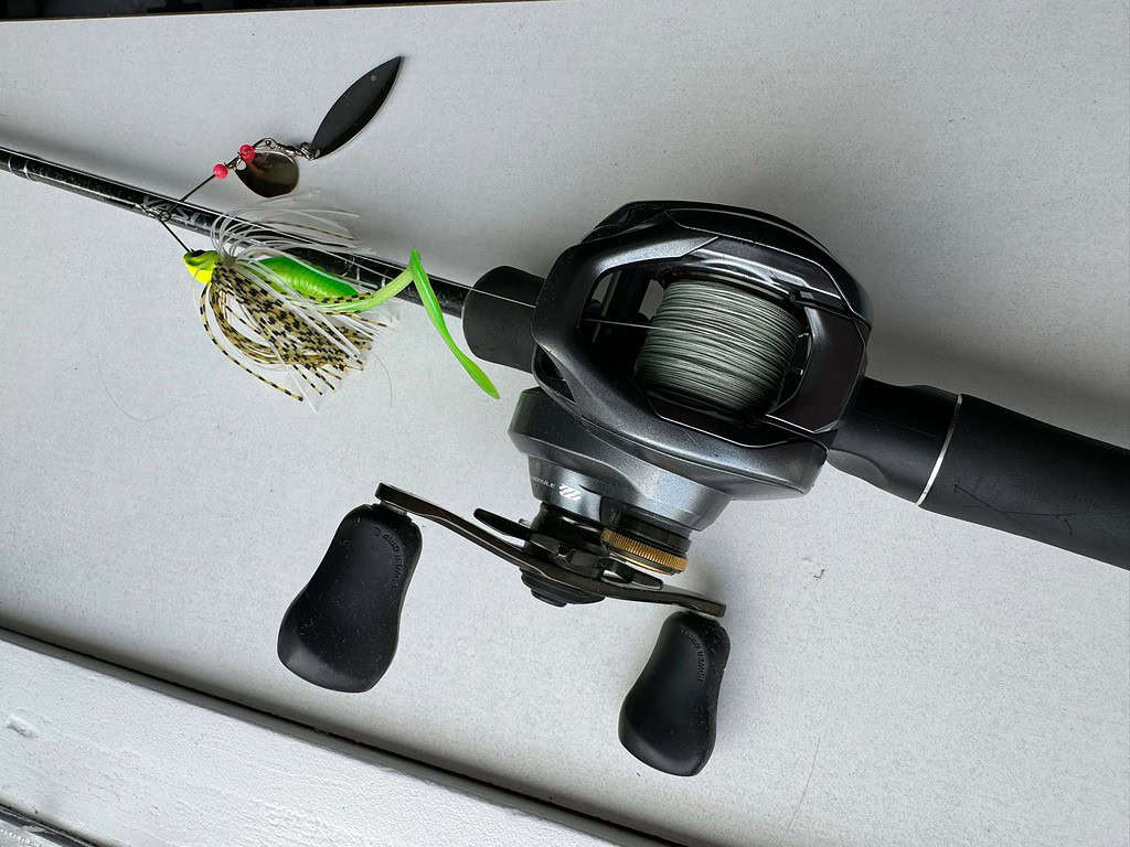 Een zwarte baitcasting-vismolen bevestigd aan een hengel met een lokaas met een groene softbait en verenrok, klaar voor vissen op baars. De uitrusting is zorgvuldig gerangschikt op een plat, wit oppervlak, klaar voor uw volgende avontuur.