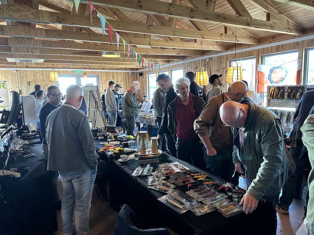 Mensen snuffelen rond op een drukbezocht indoor marktevenement op 29 september 2024. Houten balken zijn zichtbaar aan het plafond en natuurlijk licht komt binnen via de ramen, waardoor de verschillende items die worden tentoongesteld worden verlicht. Het voelt als een gezellige reünie, waarbij iedereen onder één dak samenkomt voor een unieke Roofvis Experience.