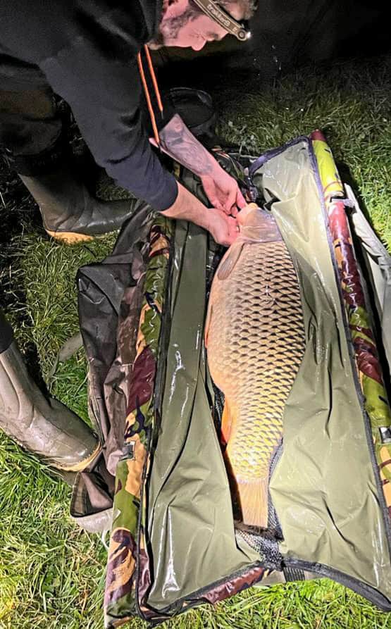 Een persoon bevestigt een karper op een gevoerde mat buiten 's nachts bij een groot water. Met een hoofdlamp, laarzen en een shirt met lange mouwen, behandelt de visser voorzichtig de vis met zijn lichtgekleurde lichaam en schubbenpatroon.