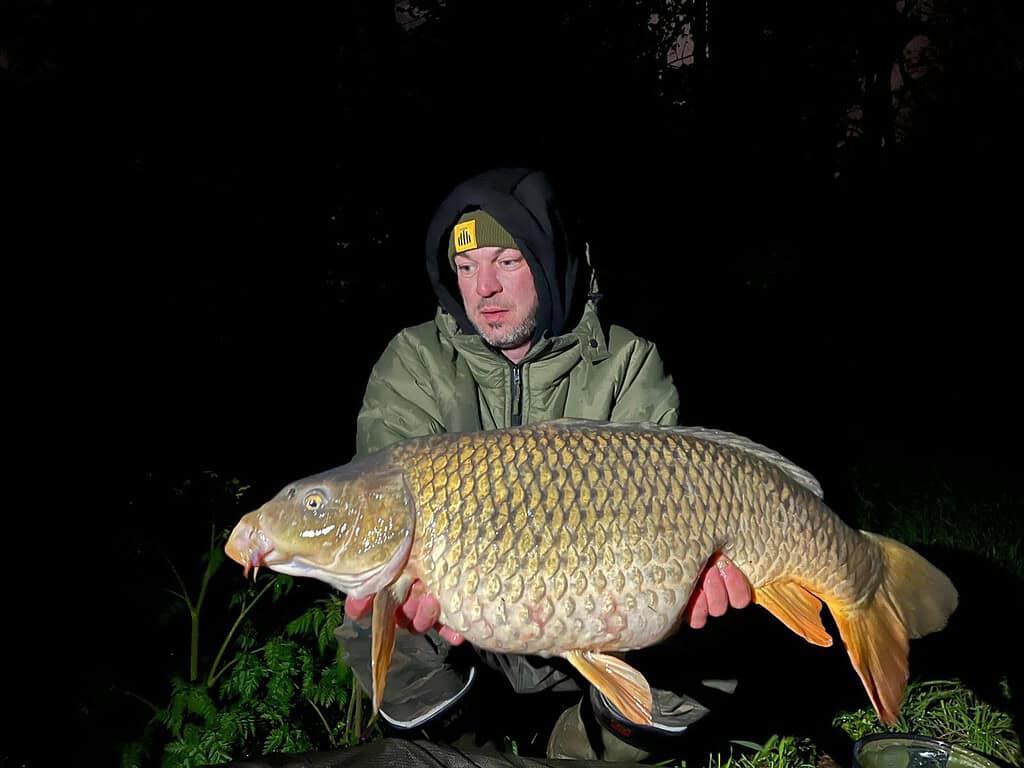 Een persoon in outdoorkleding houdt een grote karper vast en stelt deze 's nachts tentoon aan een groot water, wat de aanpak van Johan van Ingen benadrukt.