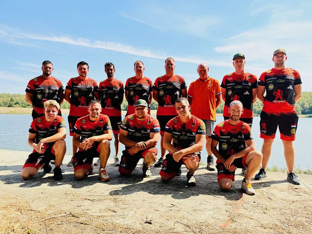 Een groep van twaalf mensen in bijpassende rood-zwarte sportkleding, gecoördineerd door Cindy Horstman, poseert buiten op een zonnige dag met water en bomen op de achtergrond.