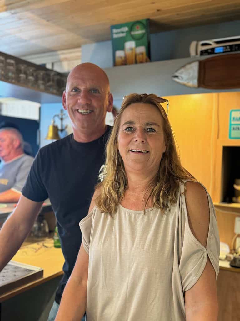 Twee mensen lachen in een keuken; de een is een kale man in een zwart shirt, de ander is een vrouw met lang blond haar in een lichtgekleurd topje.