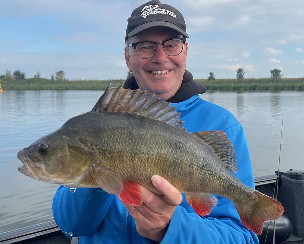 Een persoon in een blauw jasje en hoed lacht terwijl hij een grote vis met stekelige rugvinnen en rode vinnen vasthoudt, waarmee hij zijn vangsten laat zien in het kalme water.