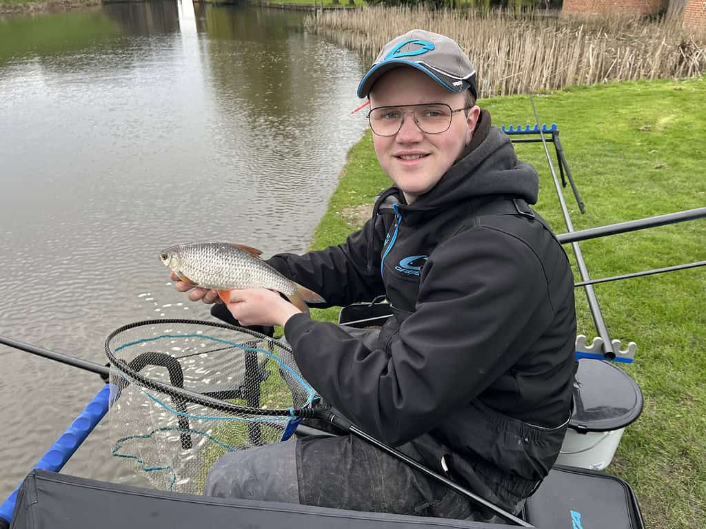 Een persoon met bril en visgerei zit aan een kalm water en houdt een vis boven een net. Achter is wat groen gras en hoog riet, wat doet denken aan een NK Dobbervissen verslag van Jelle Zwijnenberg.