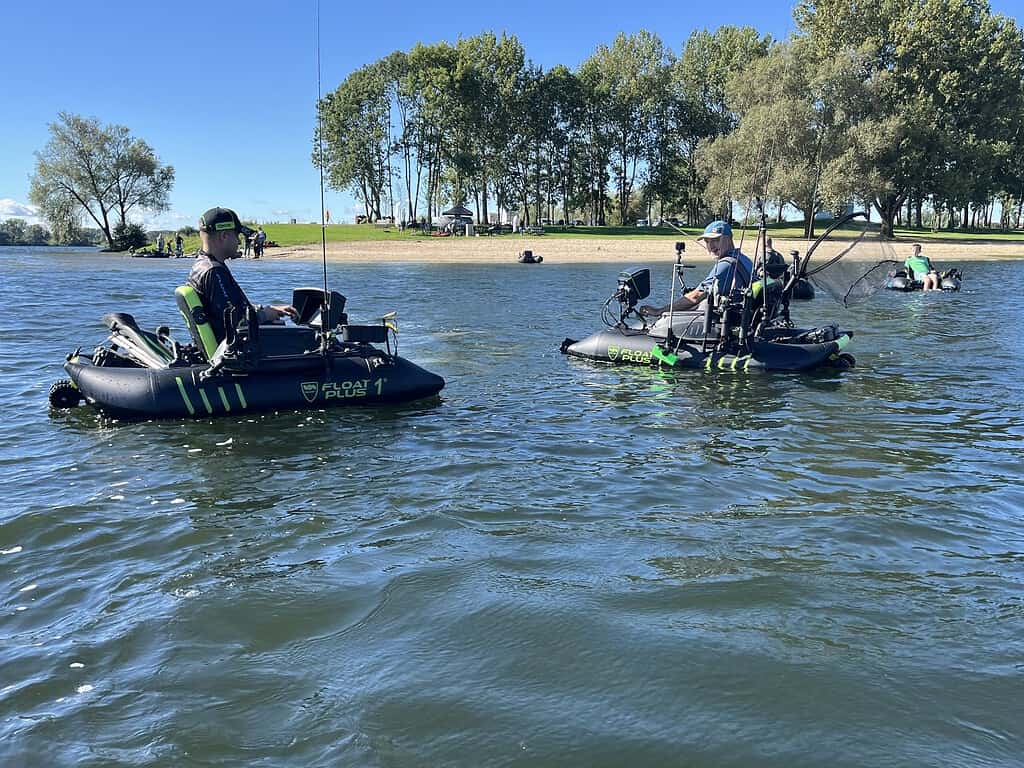 Twee mensen genieten van een Bellyboat Experience, vissend vanuit hun eigen opblaasbare boten op een kalm meer. De serene omgeving van Zandmeren heeft bomen en een grasachtige oever op de achtergrond.