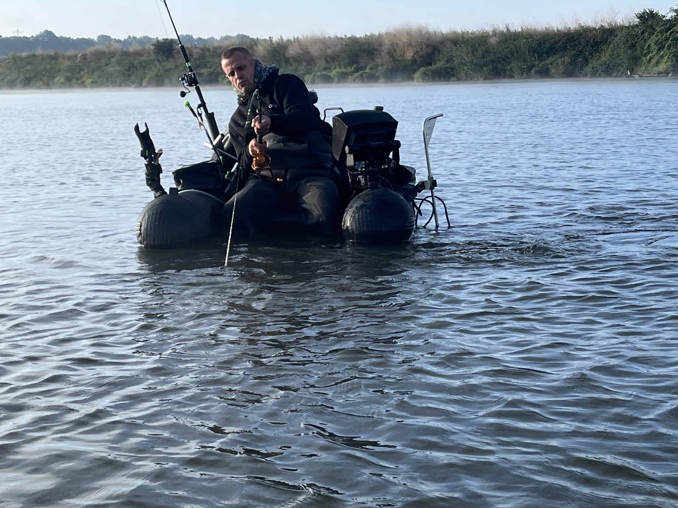 Een persoon zit rustig op een klein drijvend platform met visgerei, midden in een kalm wateroppervlak met een zanderige kustlijn op de achtergrond.