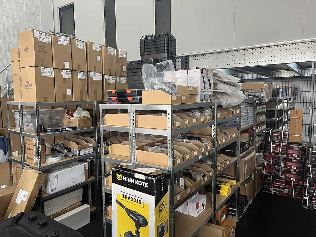 Metalen planken in een nieuw pand zijn gevuld met verschillende dozen en pakketten, waaronder producten met het label Minn Kota en Traxxis van Visboot Totaal. Andere kartonnen dozen en ingepakte artikelen zijn bovenop en in de buurt gestapeld.