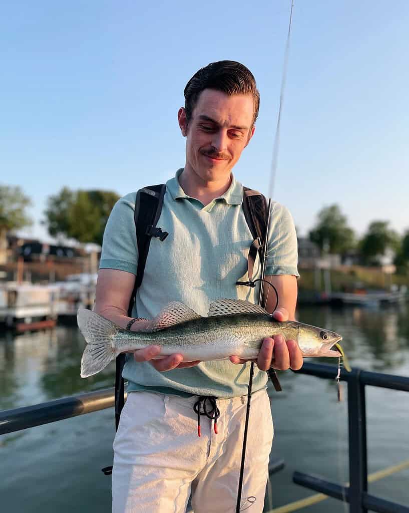 Tom Huiberts staat op een steiger met een vers gevangen vis in zijn hand en een hengel, genietend van een dagje snoekbaarsvissen. De serene achtergrond bestaat uit de rivier, bomen en gebouwen.