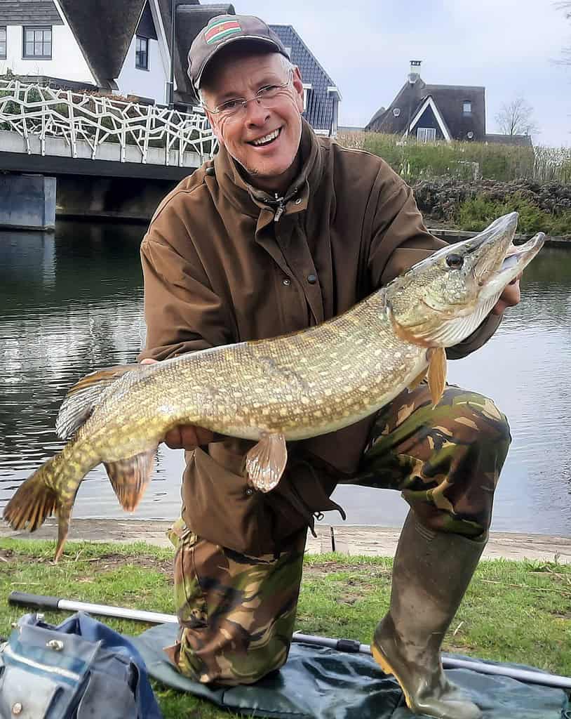 Persoon knielt bij een rivier, draagt outdoorkleding en houdt een grote vis vast. Op de achtergrond zijn een brug en huizen te zien.