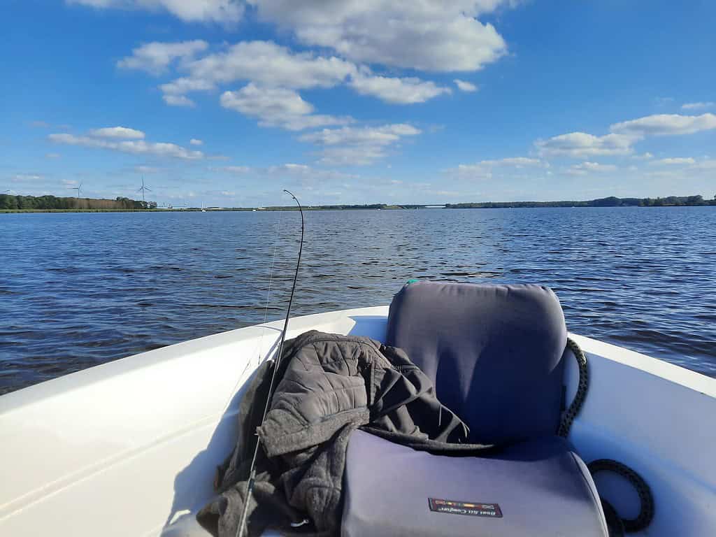 Vanaf een boot op een kalm meer ontvouwt zich het uitzicht onder een blauwe lucht met verspreide wolken. Op de voorgrond, naast een paar laarzen, rust een grijze stoel en jas. De rust belooft een ideale vissessie die zelfs Krijn van Urk zou koesteren.