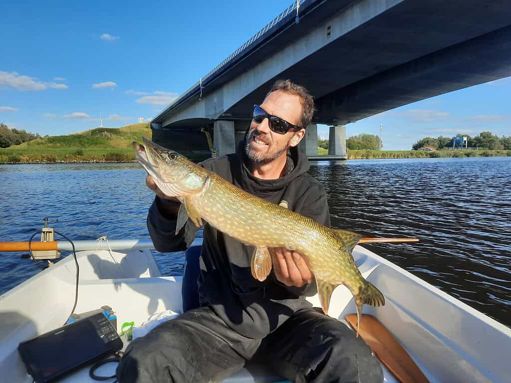 Krijn van Urk, midden in een memorabele vissessie, toont trots een grote vis terwijl hij in zijn boot onder een brug zit. Het weer is perfect, met een strakblauwe lucht die het serene moment versterkt.