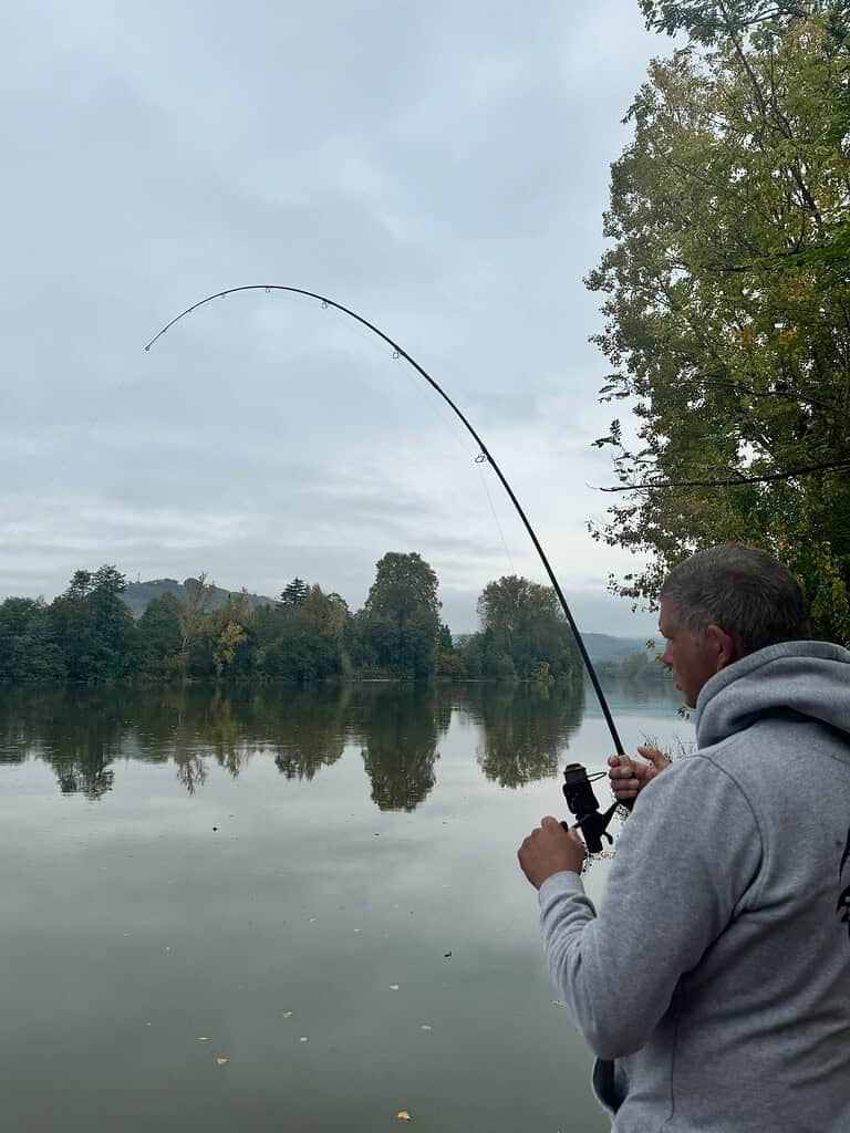 Cindy Horstman, in een grijze hoodie, vist bij een kalme, met bomen omzoomde rivier. Ze houdt een kromme hengel vast in de hoop barbeel te vangen onder de bewolkte lucht erboven.