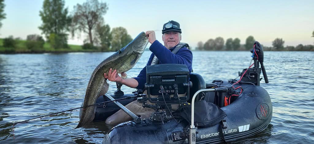 Een persoon in een kleine opblaasbare boot houdt een grote vangsten omhoog op een kalm meer, omlijst door de serene mix van bomen op de achtergrond.