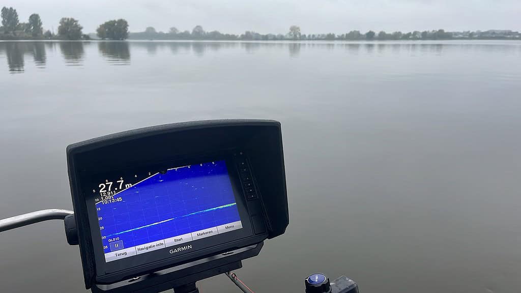 Het fishfinderscherm geeft een diepte van 27,1 meter aan op een kalm meer, met bomen op de achtergrond. Zo komt u nog dichterbij de serene schoonheid van de natuur.