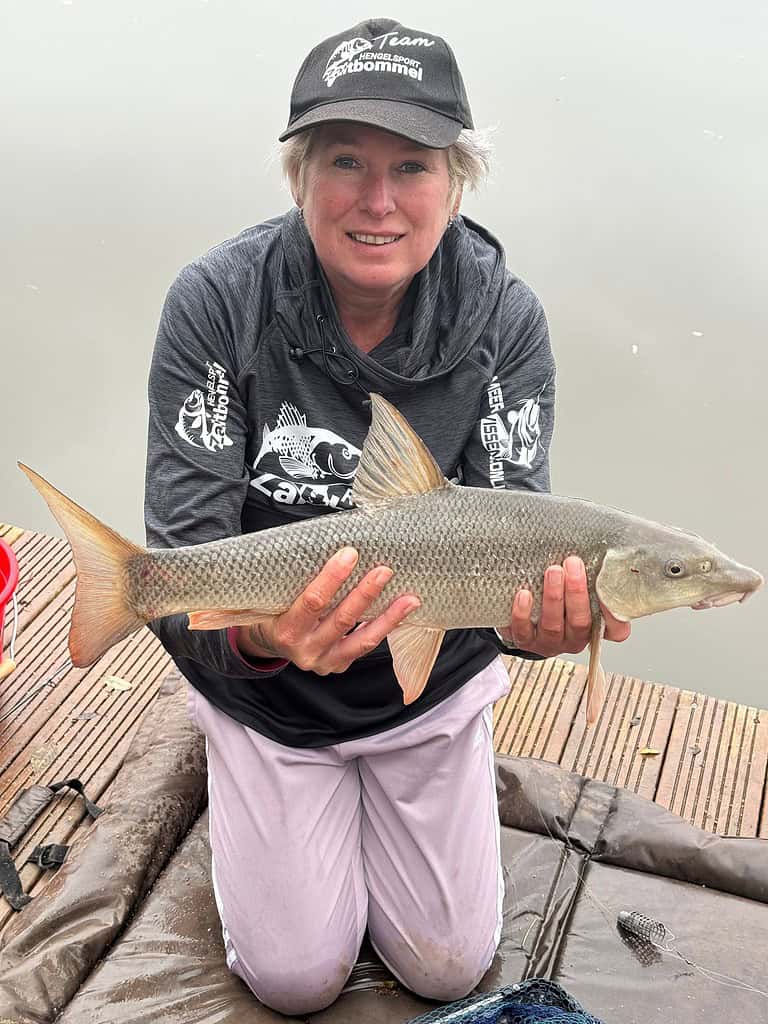 Cindy Horstman knielt op de kade, met trots een grote barbeel in haar handen. Ze draagt een pet en een donker shirt met lange mouwen, met een uitgestrekt wateroppervlak achter haar.