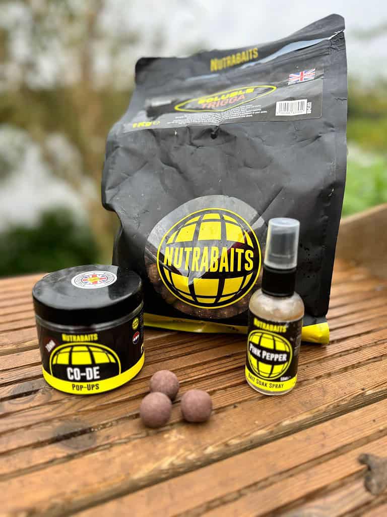 De visvoerproducten van Nutrabaits, vakkundig samengesteld voor visliefhebbers, bestaan uit een grote tas, een verpakking met pop-ups, een spray om het aas in te weken en drie ronde aassoorten die op een houten oppervlak buiten zijn uitgestald.