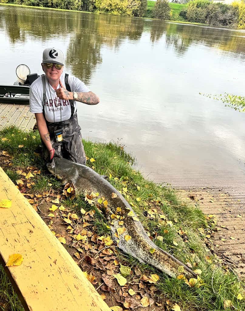 Een persoon knielt naast een grote vis bij een meer, met bomen op de achtergrond.