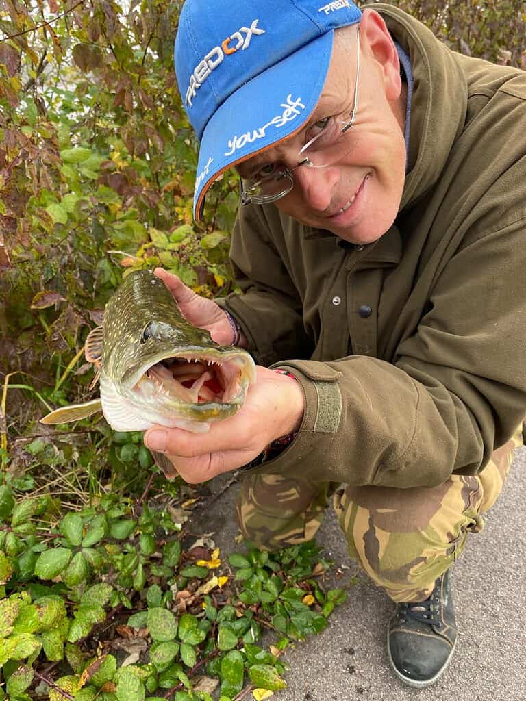 Een persoon met een pet en een bril houdt een vis vast met zijn bek open. Hij staat op een pad omgeven door groen.