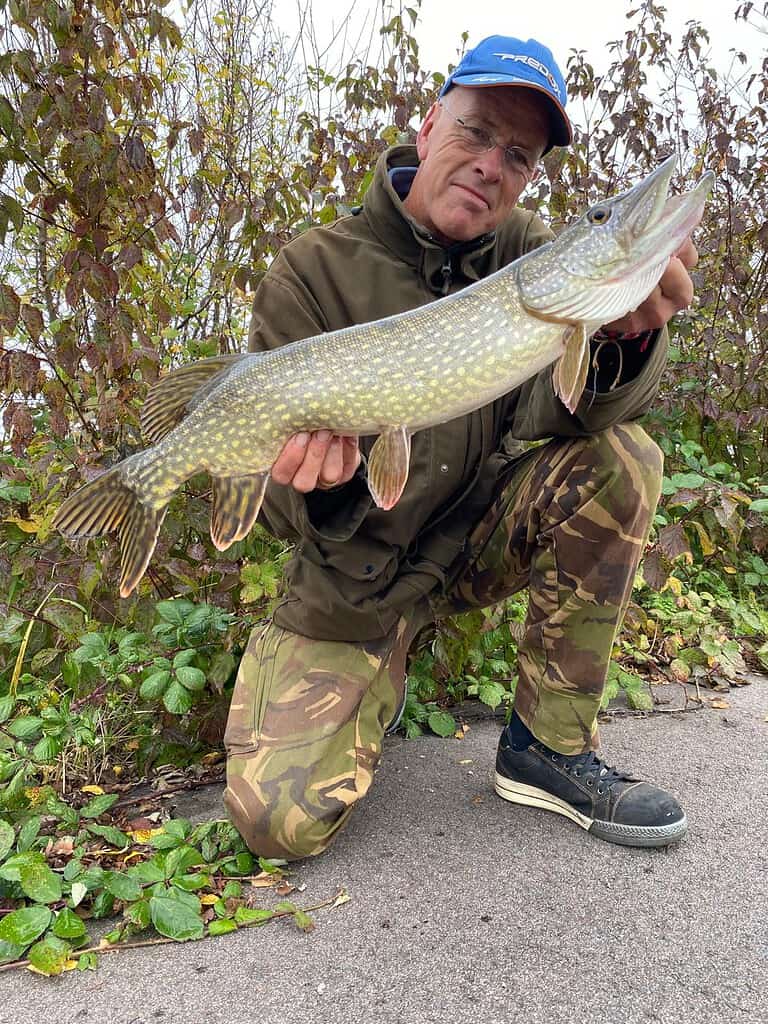 Krijn van Urk knielt buiten, trots een grote vis vasthoudend die hij heeft gevangen tijdens het snoekvissen. Met een blauwe pet, bril, een donkergroen jasje en een camouflagebroek belichaamt hij de geest van een fervent visser.