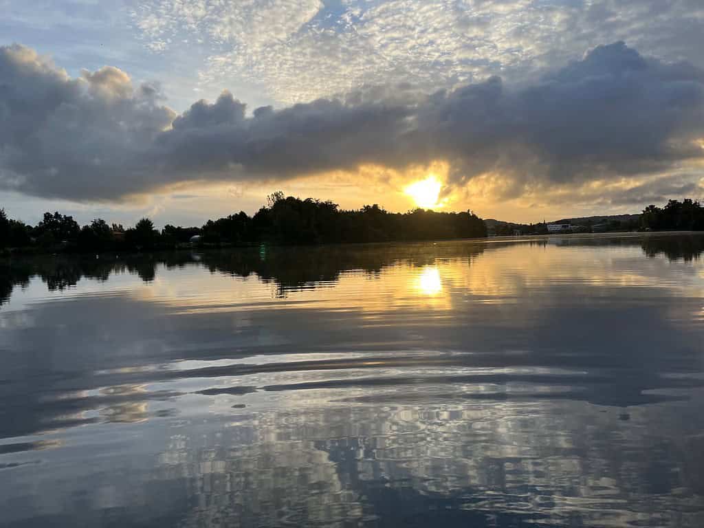 Zonsopgang boven een rustig meer in de Lot-regio in Frankrijk, met silhouetten van bomen aan de horizon en wolken in de lucht die worden weerspiegeld in het water. Dit creëert een serene achtergrond voor iedereen die op zoek is naar een ochtendavontuur waarbij u op zwarte baars vist.