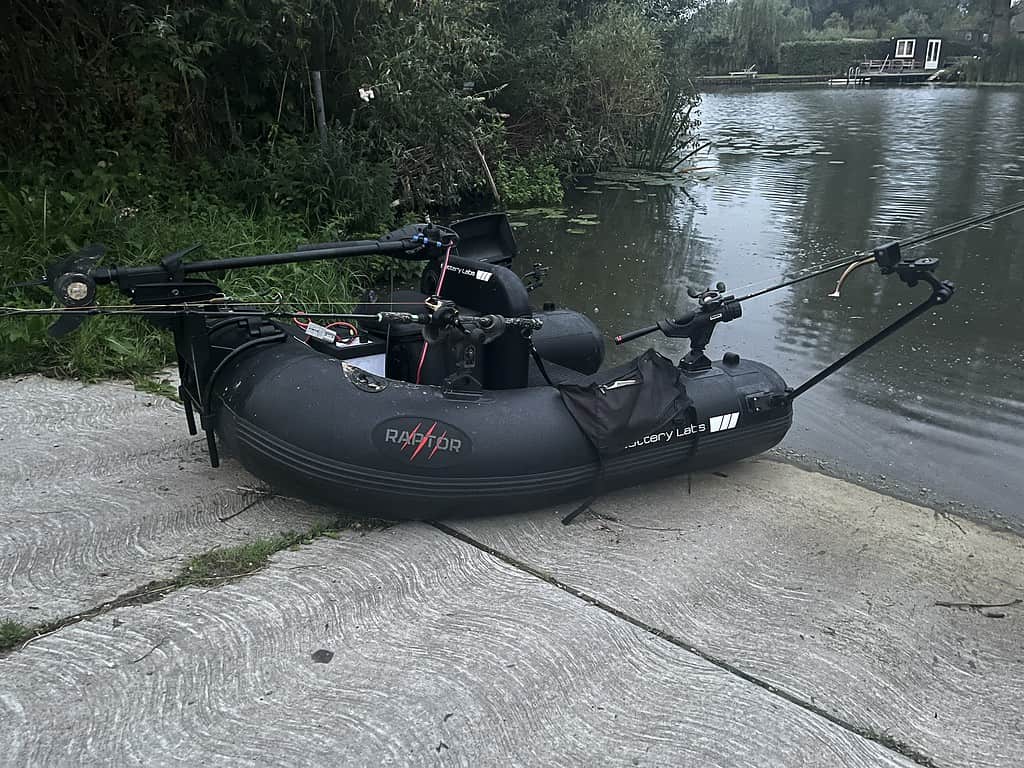 Een zwarte opblaasbare vissersboot met twee gemonteerde hengels ligt dichterbij dan ooit op een betonnen pad bij een vijver, omgeven door weelderig groen.