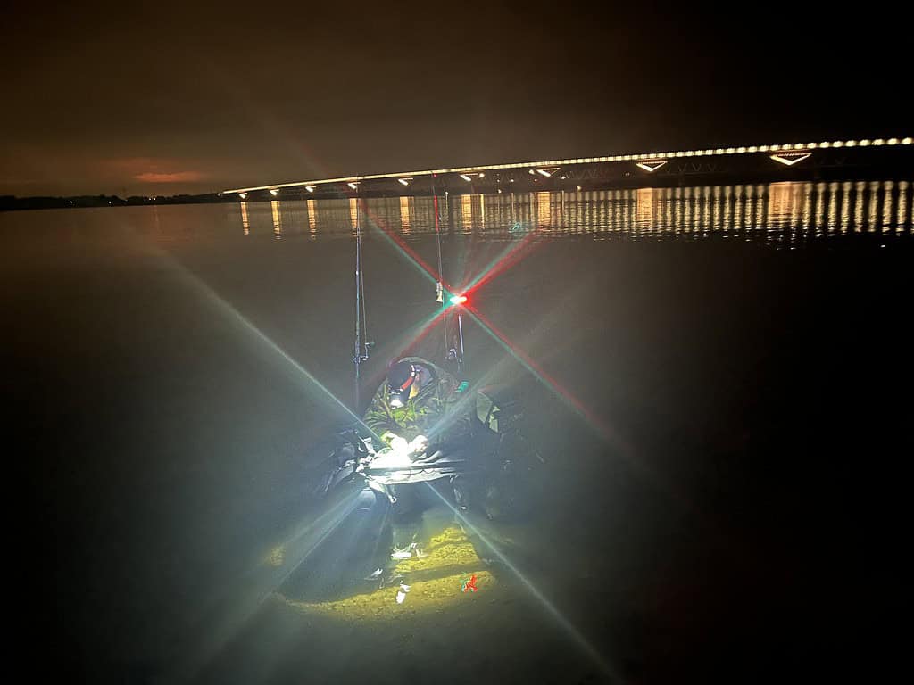 Een goed verlichte kajak op een donker meer, vlakbij een fel verlichte brug in de nacht.