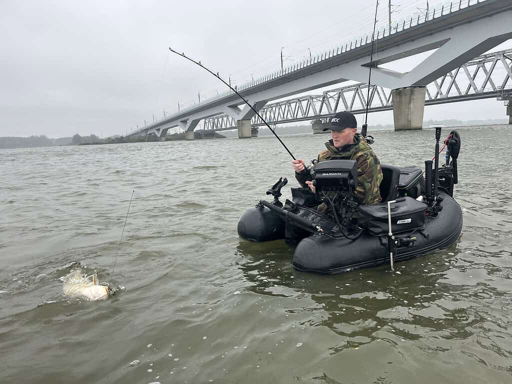 Een persoon in een kleine opblaasbare boot vangt een vis met behulp van een hengel in een rivier vlak bij een grote brug.