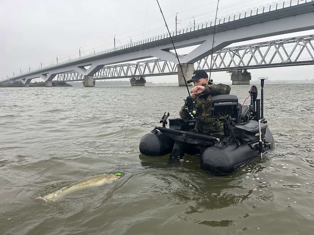 Een persoon in camouflagekleding zit op een opblaasbare boot te vissen in een watermassa bij een lange brug. Een grote vis is zichtbaar bij het wateroppervlak.