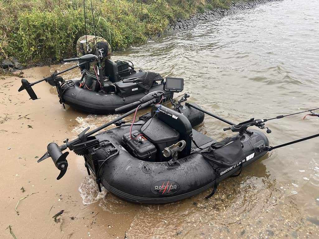 Twee opblaasbare zwarte vissersboten, uitgerust met elektromotoren, navigatieschermen en hengels, liggen voor anker op een zanderige oever vlak bij de waterkant.